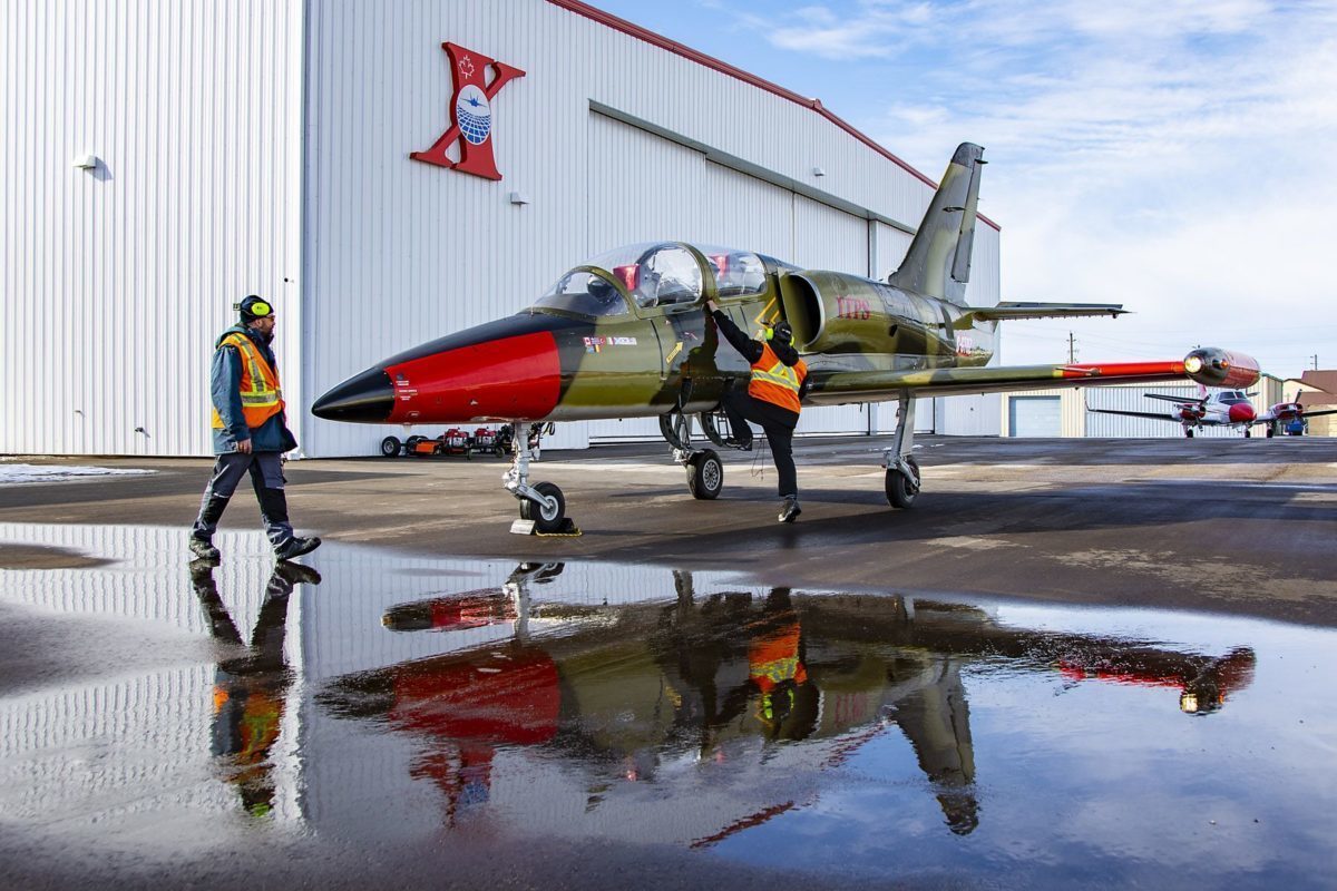 A red and green-coloured Aero Vodochody L-39C military aircraft parked with two grounds crew personnel inspecting the aircraft.