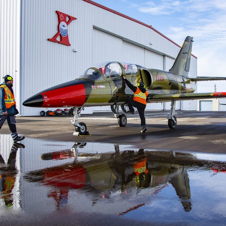 A red and green-coloured Aero Vodochody L-39C military aircraft parked with two grounds crew personnel inspecting the aircraft.
