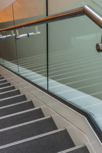 Stairs are leading downward to a small platform. The stair steps are gray. A metal handrail and glass panels are next to the stairs.