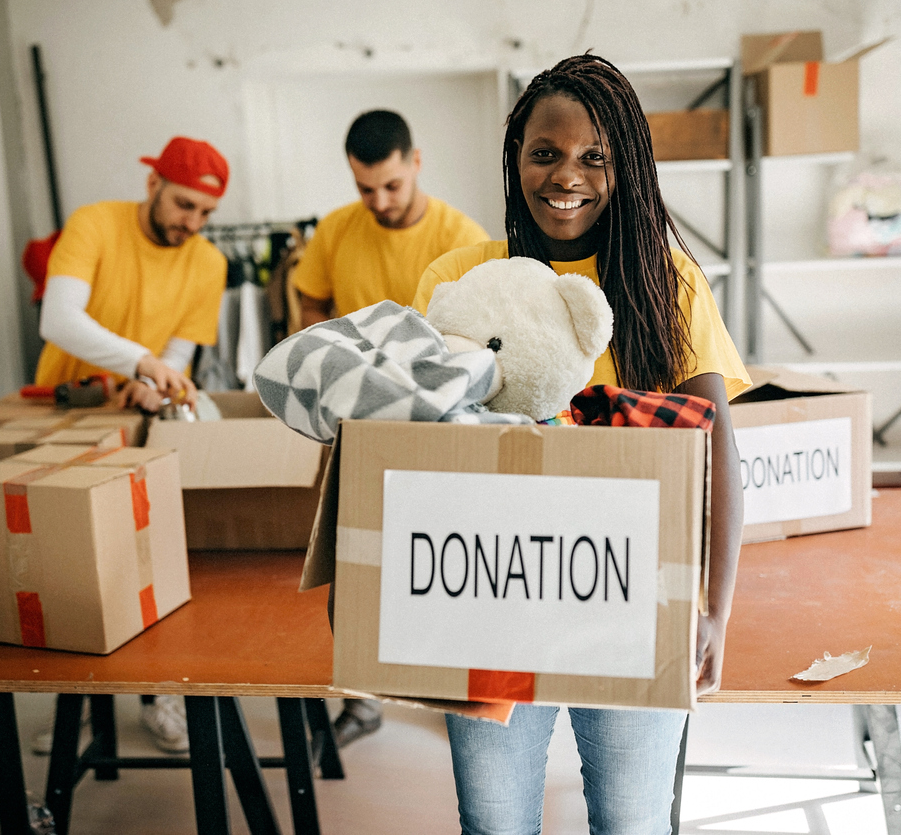 A smiling person carries a box of donated stuff including a teddy bear and a few blankets