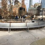 Round perforated steel bench surrounds a planting area in a park. People walk by with strollers in the background