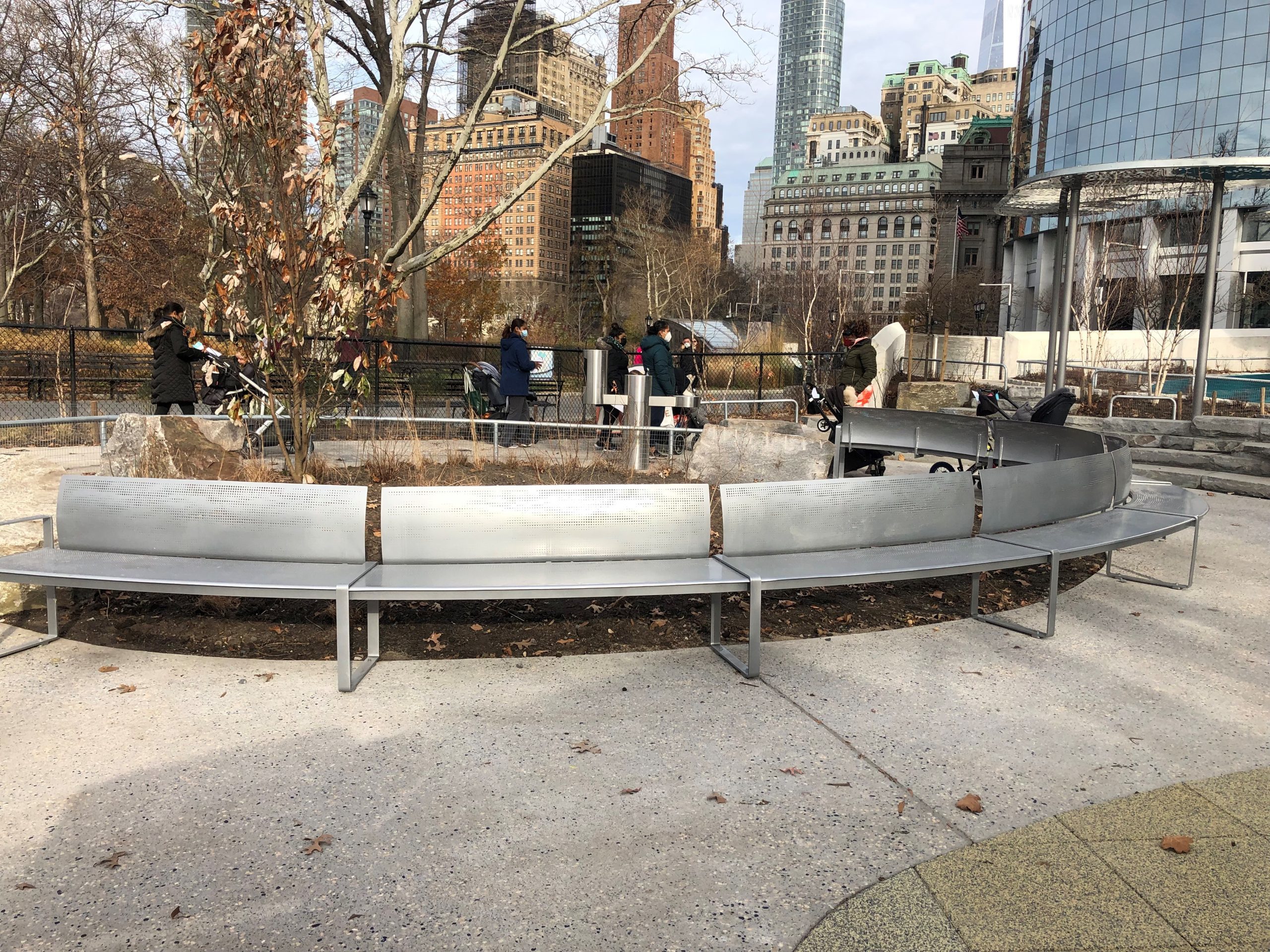 Round perforated steel bench surrounds a planting area in a park. People walk by with strollers in the background