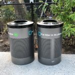 Two metal trash containers in perforated steel stand in front of a planted garden and fence
