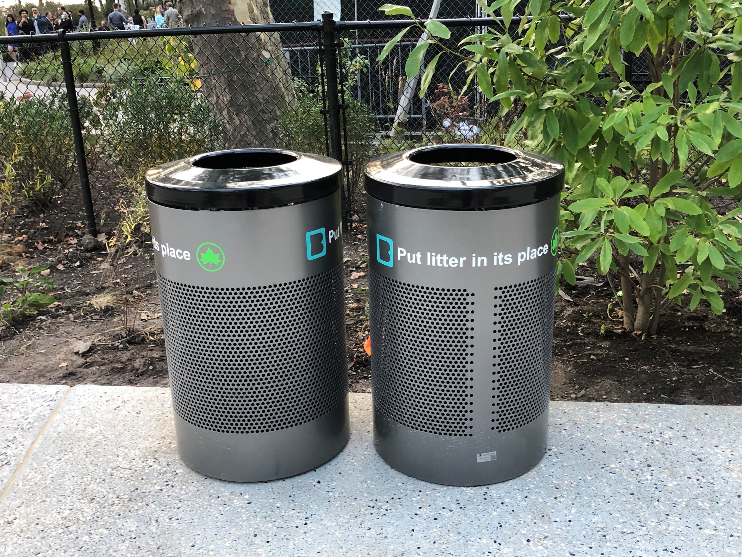 Two metal trash containers in perforated steel stand in front of a planted garden and fence