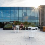 Two women chat while sitting on white Foro Bar height stools at a table in front of a large planter box