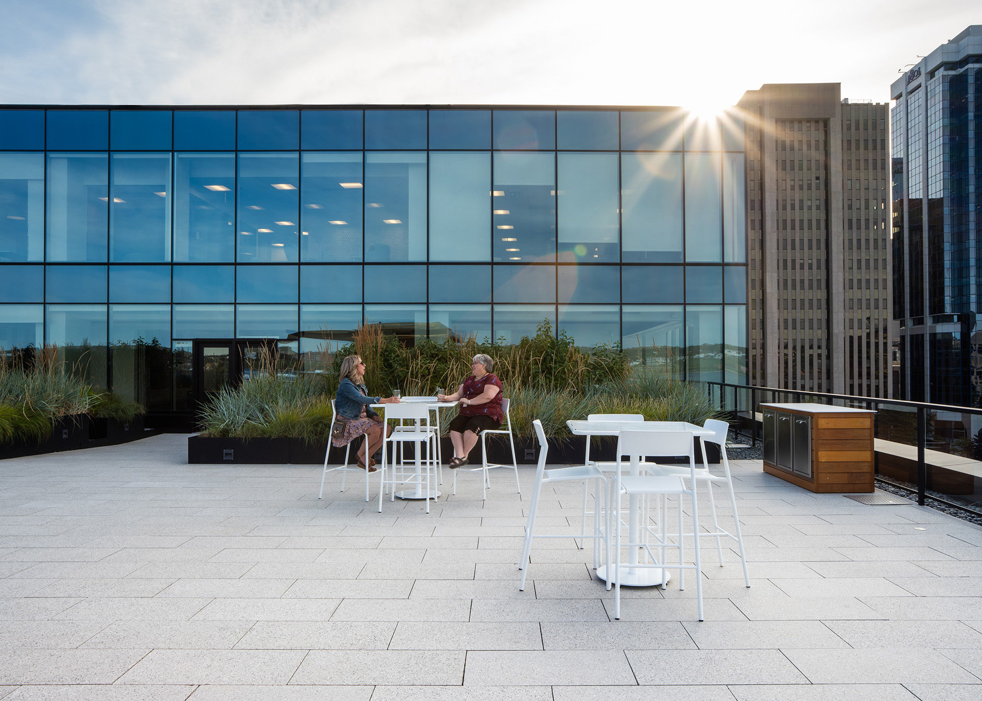 Two women chat while sitting on white Foro Bar height stools at a table in front of a large planter box
