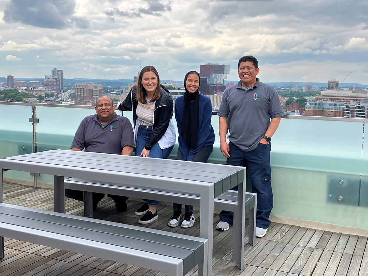 Four maintenance workers at the Viridian Apartments smile pose on the roof deck with newly installed 1050 tables and benches. The Boston skyline is in the background.