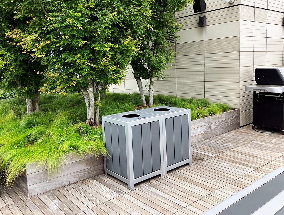 Two side-by-side 1050 Waste & Recycling containers in front of a raised planting bed with grasses and trees on the rooftop deck of the Viridian Apartments in Boston.