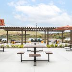 Six Maglin 400 Series Cluster Seating units are placed throughout a concrete amenity courtyard at an apartment complex in Chula Vista, CA. There is a shade pergola in the background and two striped orange and red umbrellas providing shade.