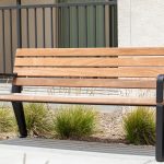 A Maglin Iconic Collection bench with wood slats and black cast aluminum end arms sits in front of a stucco apartment building. Drought-tolerant grasses are planted behind the bench in a mulched bed.