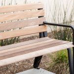A partial view of the Maglin Iconic Collection bench with wood slats and black cast aluminum end arms sits in front of a stucco apartment building. Drought-tolerant grasses are planted behind the bench in a mulched bed.