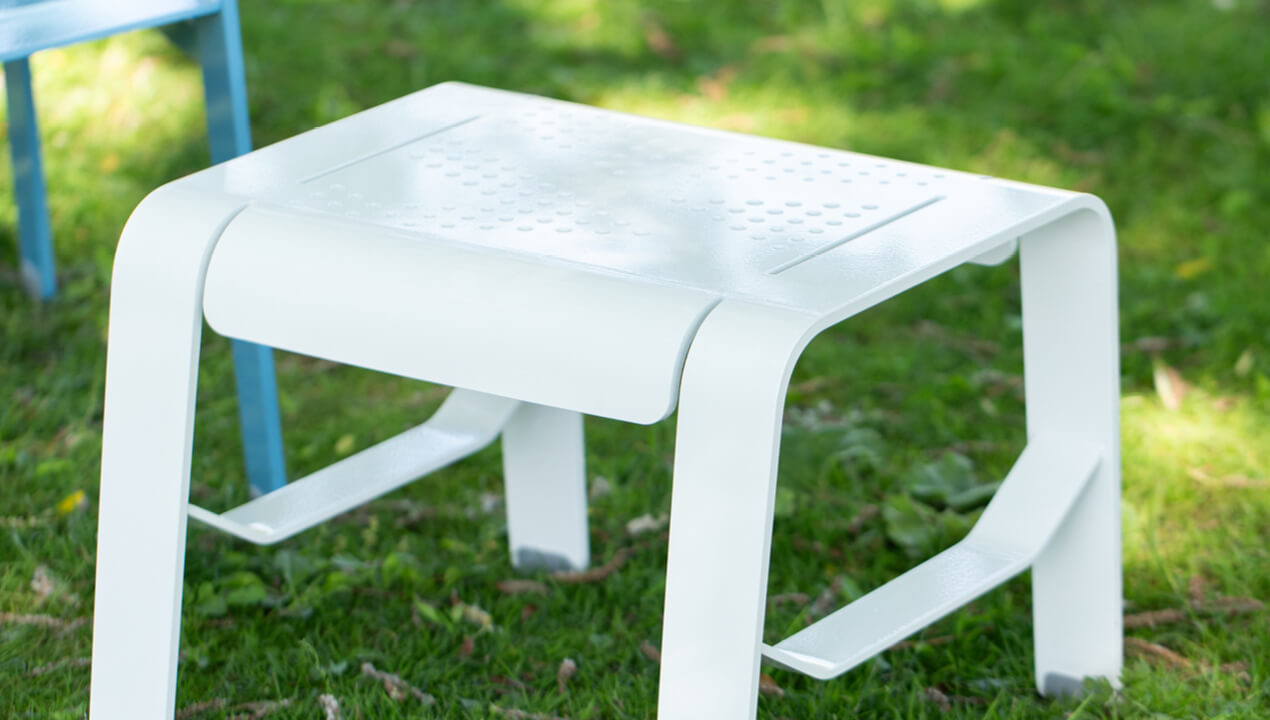 ALUM footrest/side table with Prosecco pattern in powdercoat white paint against greenery