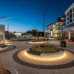 Concrete circular planters are topped with backless Ogden Benches in AGORA Plaza, Gatineau. The image is at twilight and the bench and concrete planters are underlit, casting a glow on surrounding pavement.