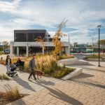 A child sits on a Maglin Ogden Bench while a woman walks by with a stroller and a man walks a poodle-type dog on a pathway in Agora Plaza, Gatineau, QC. There is plentiful outdoor seating for comfortable use outdoors.