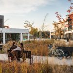 A couple sits on a Maglin Ogden Bench on a paved pathway in AGORA Plaza in Gatineau. There bikes are resting against another Ogden Bench across the path from where they sit.