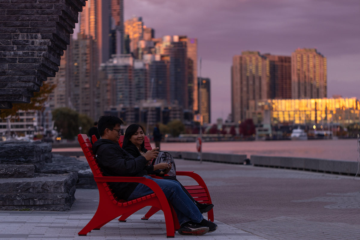 Two people sit next to each other in Maglin 720 Chairs at Bathurst Quay Common. In the twilight, the Toronto downtown skyline appears in the background. appears