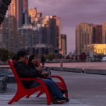 Two people sit next to each other in Maglin 720 Chairs at Bathurst Quay Common. In the twilight, the Toronto downtown skyline appears in the background. appears