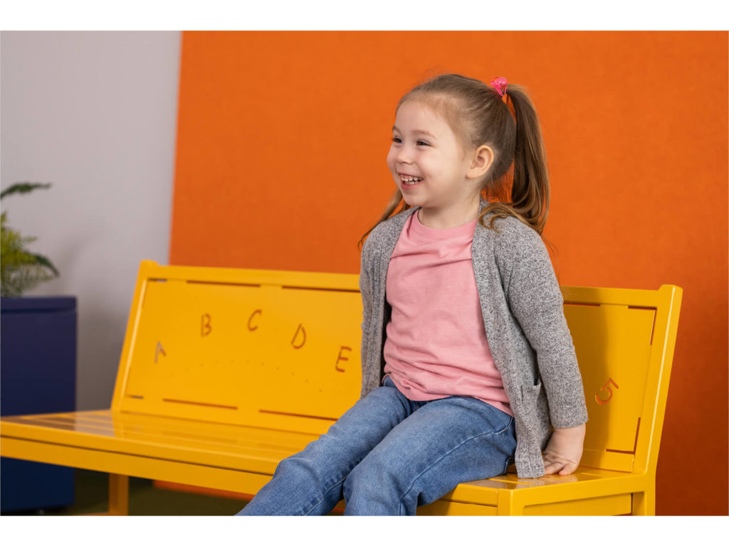Child sitting on yellow backed bench with letters against orange wall
