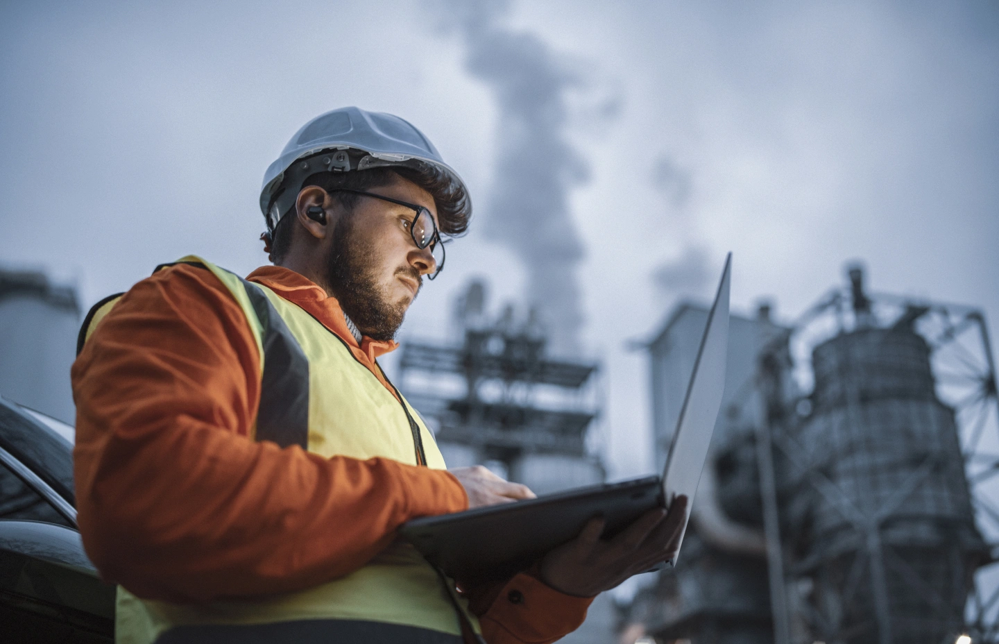 A construction worker wearing a white hard hat, glasses, and a high-visibility vest works on a laptop outdoors at an industrial site with smoke stacks in the background.