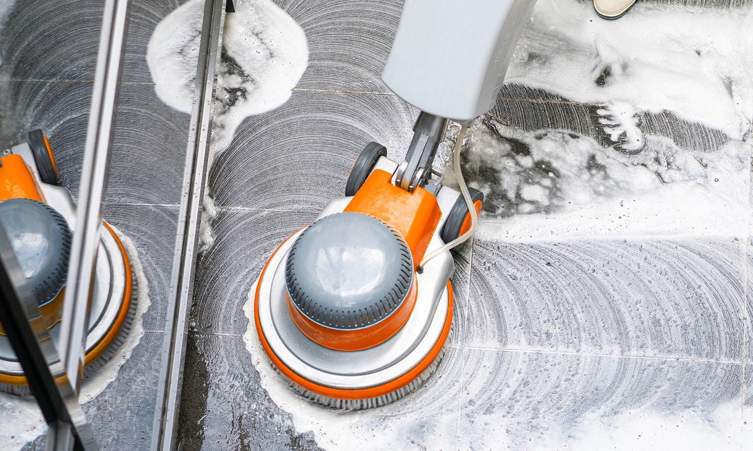 A floor-cleaning machine with a yellow base scrubs a tiled surface, leaving clean, swirling patterns amidst soapy water.