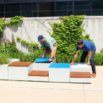 Two men assembling Pixel blocks against a wall with greenery