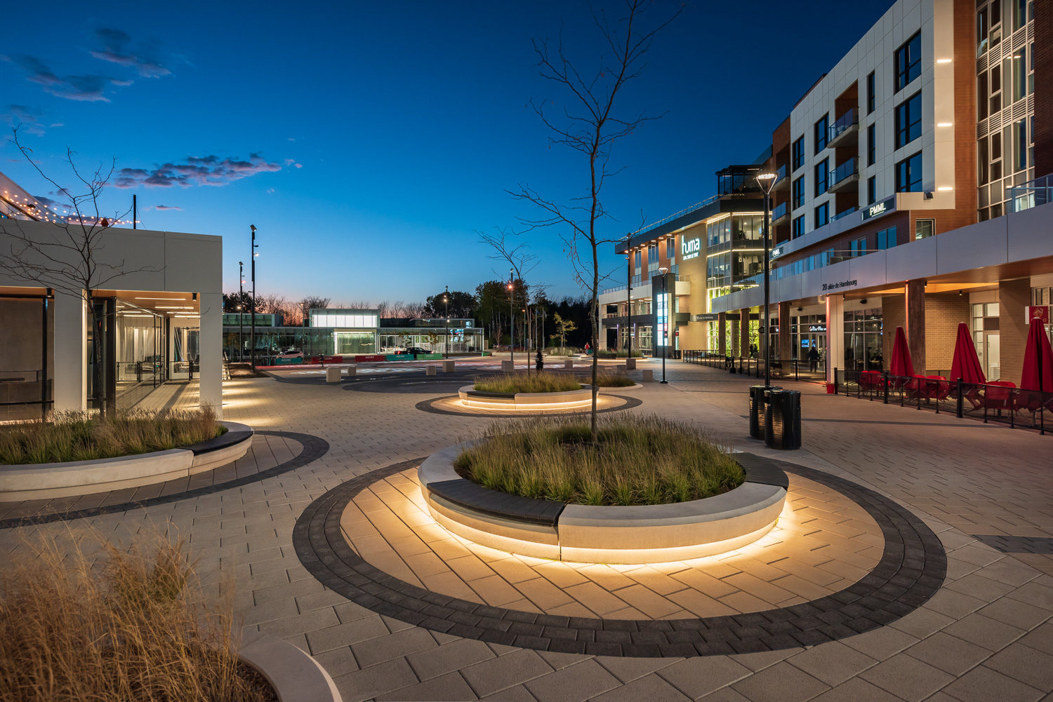 Concrete circular planters are topped with backless Ogden Benches in AGORA Plaza, Gatineau. The image is at twilight and the bench and concrete planters are underlit, casting a glow on surrounding pavement.