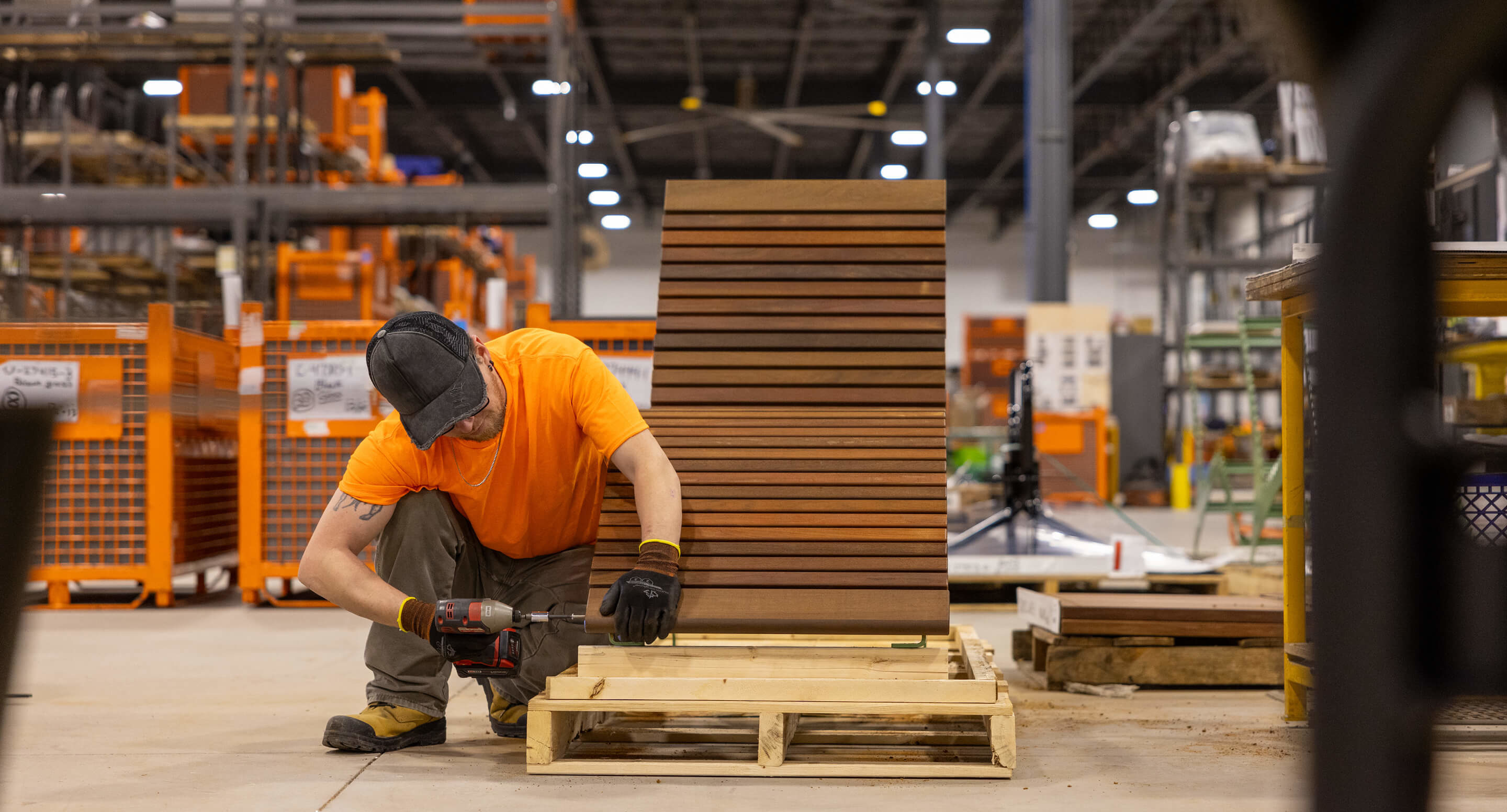 A Maglin employee wearing an orange t-shirt drills and assembles a 720 lounge chair in the warehouse.