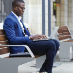 Man in blue coat sitting on bench while using laptop