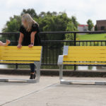 Two women chatting while one sits on backed yellow bench and other leans on it's back