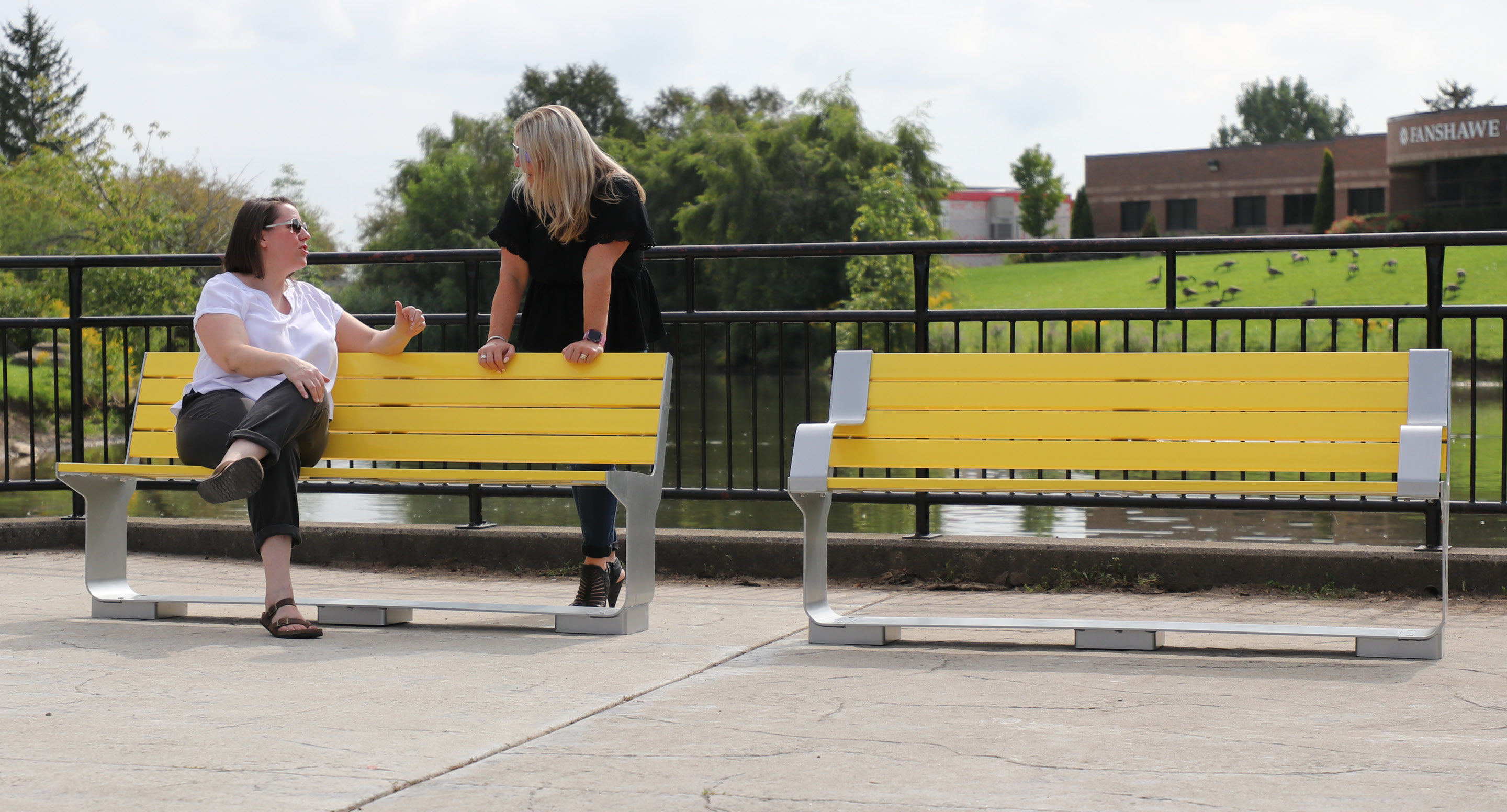 Two women chatting while one sits on backed yellow bench and other leans on it's back