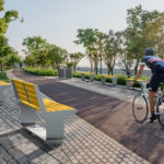 Cyclist riding past two yellow benches and women walking dog in the distance
