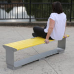 Women sitting on backless yellow L-Series bench on patio next to water and fence