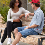 Man and woman sitting on backed wooden bench with stones and greenery in background